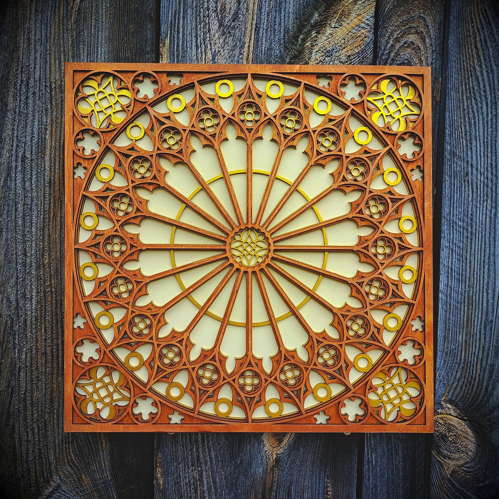 A square decorative wooden wall plaque featuring an intricate radial floral and geometric pattern in warm brown wood with gold-toned accents, displayed against a dark weathered wood plank background.